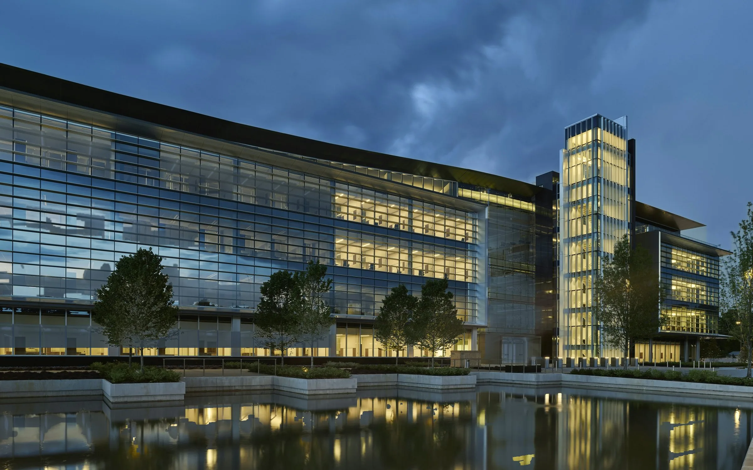 Illustrative photograph: a glass office building at dusk, its offices lit, beside a reflecting pool